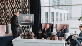 Several people are sitting together in a corner with colorfully patterned upholstered furniture, looking at a monitor on which two other people are connected.