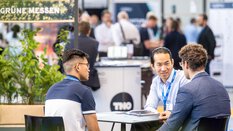 Three men sit at a table at an exhibition stand and talk.