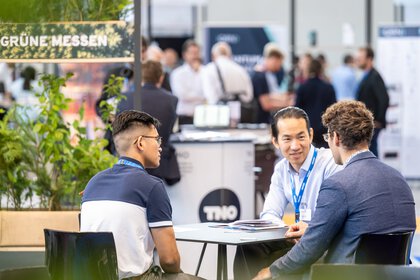 Three men sit at a table at an exhibition stand and talk.