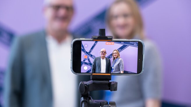 Zwei Menschen stehen vor einer Fotowand. Ein Smartphone steht vor Ihnen und filmt.