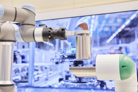A robotic arm holds a metal cylinder in a high-tech industrial environment with machines in the background.