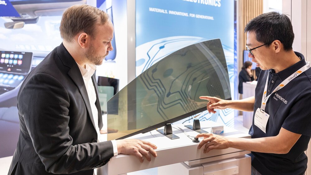 Two men are standing together at a trade fair stand at LOPEC, examining a piece of thick, flexible film more closely.