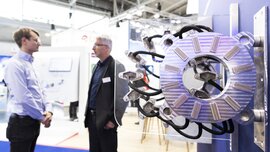 Two people talking next to a large industrial machine with cable connections on display at a trade fair stand.