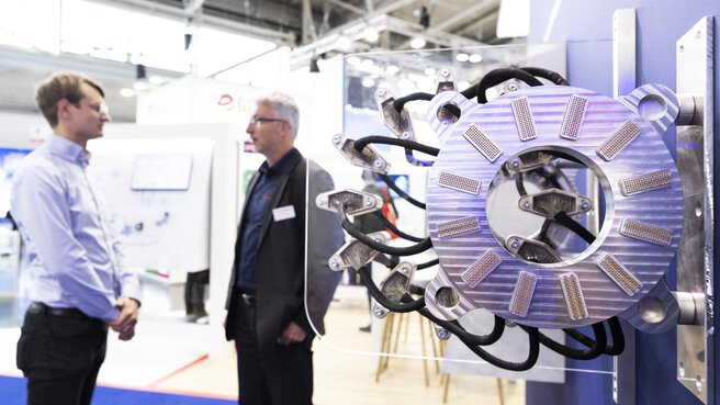 Two people talking next to a large industrial machine with cable connections on display at a trade fair stand.