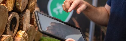 Person bedient ein Tablet an einem Messestand mit gestapelten Holzscheiben und grüner Waldbeschilderung im Hintergrund.