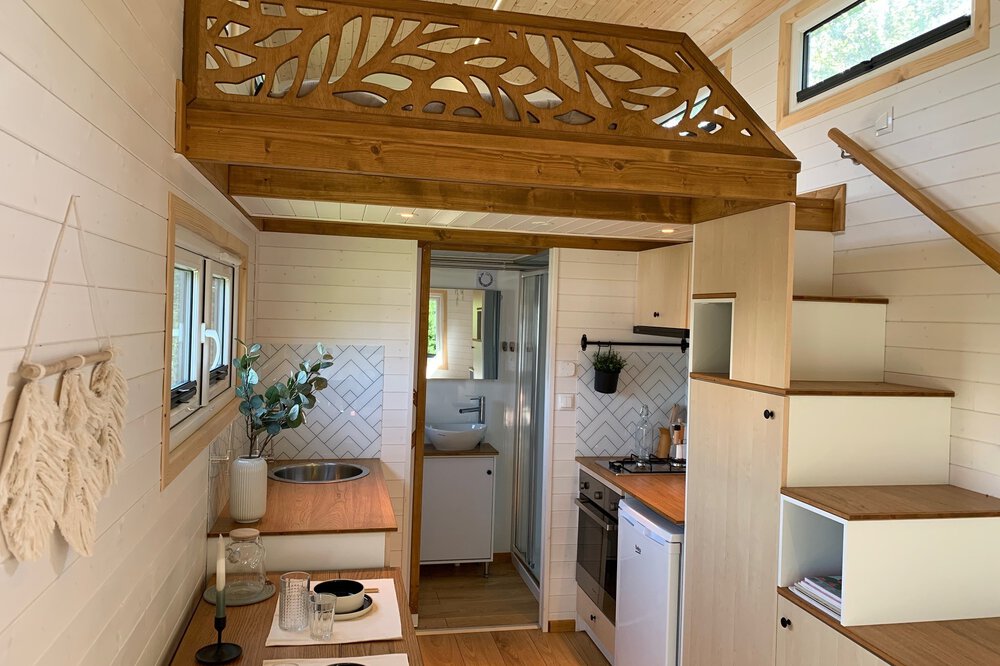 Gemütliches Tiny-House-Interieur mit Holzboden, kleiner Küche, Tisch mit Stühlen und Treppe.