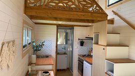 Gemütliches Tiny-House-Interieur mit Holzboden, kleiner Küche, Tisch mit Stühlen und Treppe.