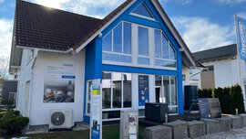 Modernes Haus mit blauen Akzenten, großen Fenstern, Sonnenkollektoren und Wärmepumpen, das im Freien unter einem sonnigen Himmel steht.