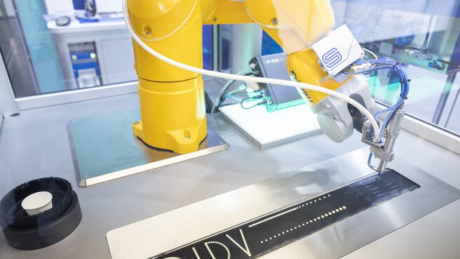 Ein gelber Roboterarm positioniert eine Art Stift über einem schwarzen Band, auf dem ein Smiley, 