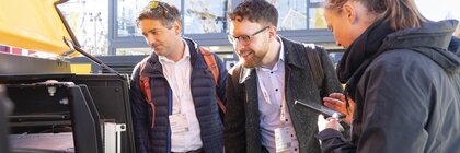 Drei Messebesucher betrachten interessiert das geöffnete Fach einer Baumaschine, eine Frau macht sich Notizen auf dem Smartphone