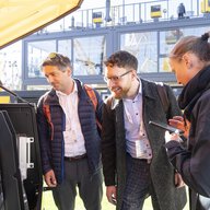 Drei Messebesucher betrachten interessiert das geöffnete Fach einer Baumaschine, eine Frau macht sich Notizen auf dem Smartphone