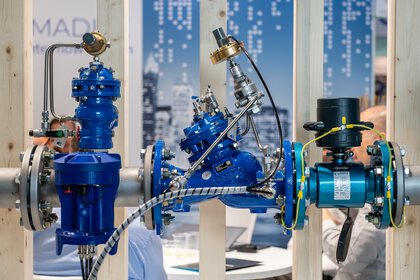 Eine komplexe blaue Industrieventilbaugruppe, montiert auf einem Ausstellungsständer mit einem Messestand im Hintergrund.