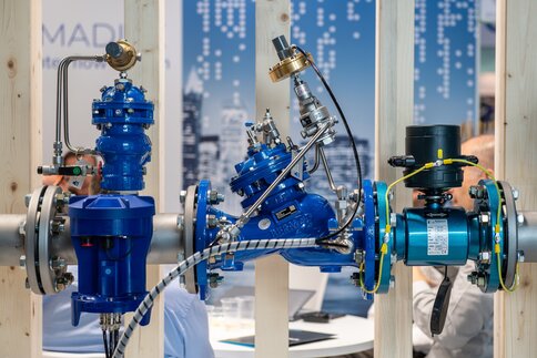 A complex blue industrial valve assembly mounted on a display stand with an exhibition stand in the background.