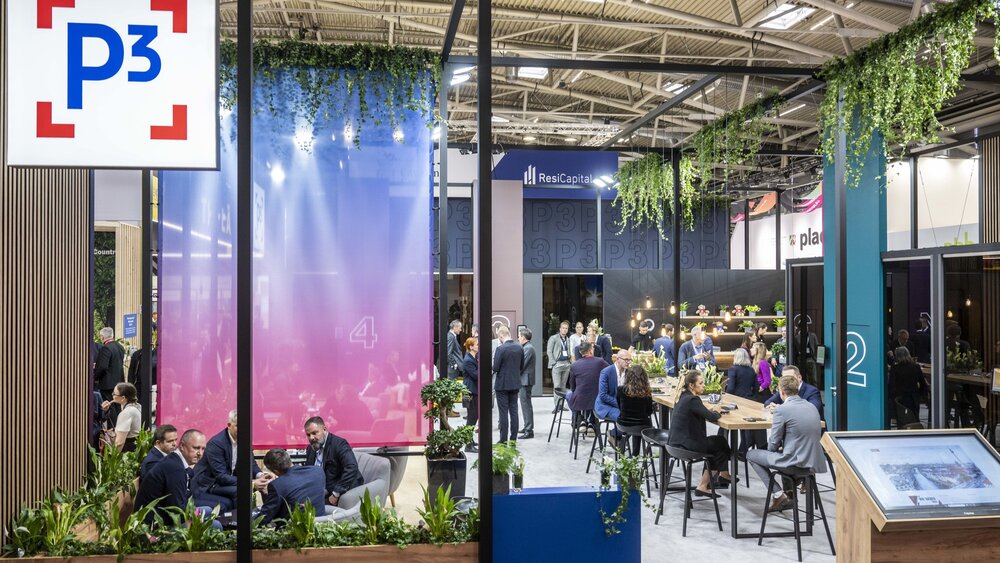 Ein Meeting-Bereich auf der EXPO REAL mit abgetrennten Sitzbereichen, Bannern, Pflanzendeko und Personen, die sich in Gruppen unterhalten.