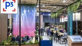 Ein Meeting-Bereich auf der EXPO REAL mit abgetrennten Sitzbereichen, Bannern, Pflanzendeko und Personen, die sich in Gruppen unterhalten.