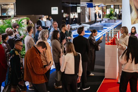 Eine große Gruppe Messebesucher steht gemeinsam vor einer Schmuckvitrine in einem Gang der ceramitec und hört dem Vortrag einer Ausstellerin zu.