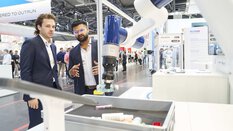Zwei Männer im Anzug stehen auf einer Messe vor einem kollaborativen Roboterarm, der ein Objekt aus einer Kiste greift. Einer der Männer erklärt gestikulierend die Technologie, während Besucher im Hintergrund durch die Ausstellung gehen.