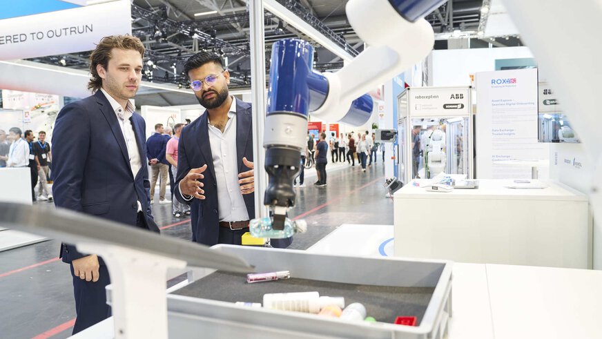 Zwei Männer im Anzug stehen auf einer Messe vor einem kollaborativen Roboterarm, der ein Objekt aus einer Kiste greift. Einer der Männer erklärt gestikulierend die Technologie, während Besucher im Hintergrund durch die Ausstellung gehen.