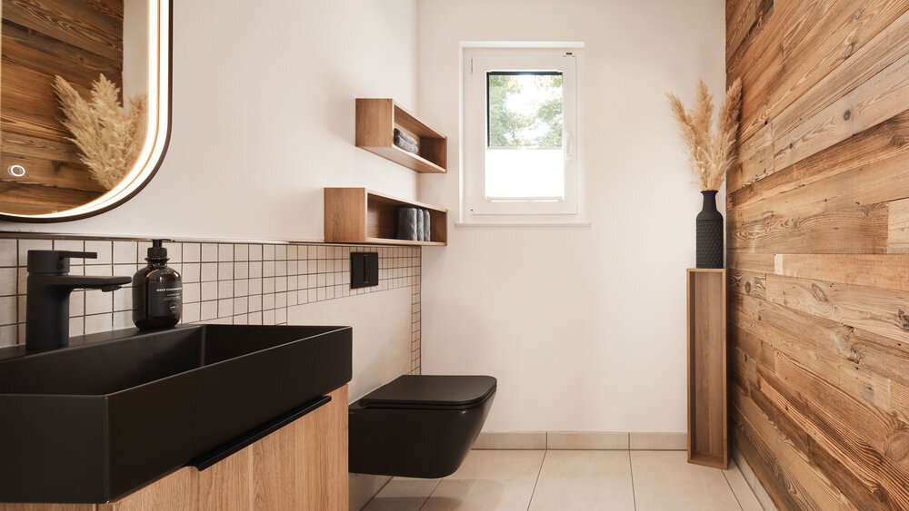Modernes Badezimmer mit Holzakzenten, schwarzen Armaturen, einer wandmontierten Toilette und einem kleinen Fenster, das Tageslicht hereinlässt.