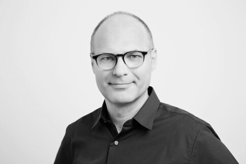 Ein Mann mit Brille in schwarzem Hemd, leicht lächelnd, vor weißem Hintergrund.