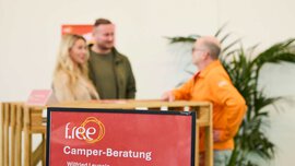 Im Fokus ein Schild mit der Aufschrift „f.re.e Camping-Beratung“ und „Wilfried Leupolz“. Dahinter stehen zwei Männer und eine Frau und unterhalten sich.