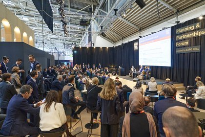 Eine gut besuchte Podiumsdiskussion im „Planning & Partnerships Forum“ der EXPO REAL in München. Zahlreiche Besucherinnen und Besucher sitzen und stehen vor der Bühne, auf der mehrere Expertinnen und Experten sprechen. Über der Bühne hängt ein großes Display mit Informationen zur Veranstaltung, und an der Seite ist das Forum mit „curated by EXPO REAL“ beschriftet. Die Atmosphäre ist konzentriert und professionell.