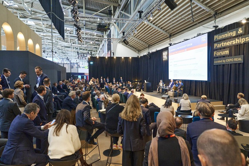 A well-attended panel discussion at the “Planning & Partnerships Forum” during EXPO REAL in Munich. Numerous attendees are seated and standing in front of the stage, where several experts are speaking. Above the stage, a large screen displays event information, and the forum signage reads “curated by EXPO REAL.” The atmosphere is focused and professional.
