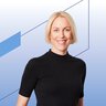 Eine lächelnde Frau mit kurzen blonden Haaren in einem schwarzen Oberteil steht vor einem blauen geometrischen Hintergrund.
