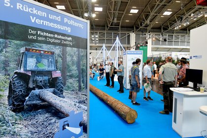 Stand with tree trunk and blue carpet, on which several visitors to INTERFORST are standing. The focus is on a poster about the 