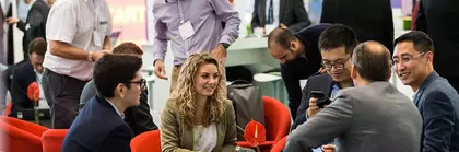 A group of trade fair visitors sit in a circle on red chairs and chat, while other visitors and exhibitors can be seen in the background.
