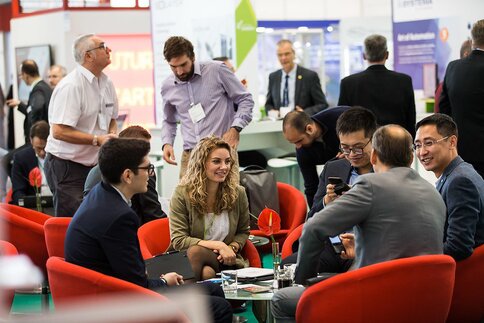 A group of trade fair visitors sit in a circle on red chairs and chat, while other visitors and exhibitors can be seen in the background.