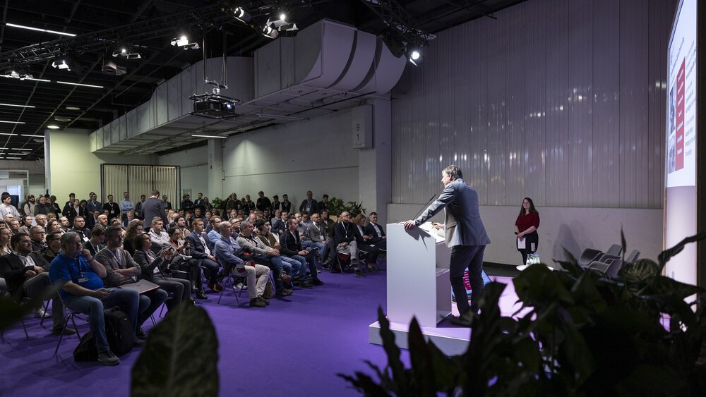 Ein Redner spricht von einem Podium aus zu einem sitzenden Publikum in einem großen, modernen Konferenzsaal mit lila Bodenbelag.