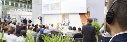 Blurred trade fair stage with seated audience, in the foreground is a visitor in a suit with headphones.