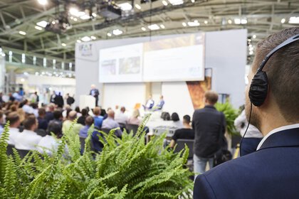 Blurred trade fair stage with seated audience, in the foreground is a visitor in a suit with headphones.