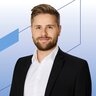 Porträtfoto eines bärtigen Mannes, der ein weißes Hemd und einen schwarzen Blazer trägt.