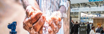 Darstellung eines Wasserleitungssystems vor einem großen Display, auf dem Hände zu sehen sind, die Wasser schöpfen, mit dem Text „Water is life“.
