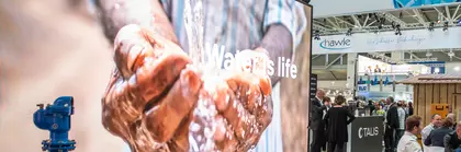 Darstellung eines Wasserleitungssystems vor einem großen Display, auf dem Hände zu sehen sind, die Wasser schöpfen, mit dem Text „Water is life“.
