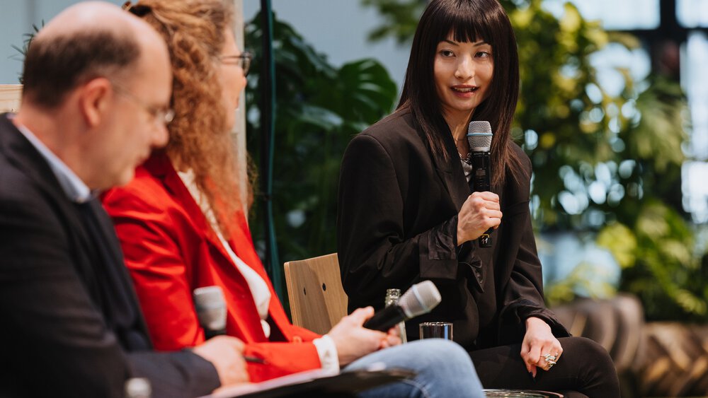 Drei Personen halten Mikrofone, sitzen und unterhalten sich in einer Diskussionssituation, im Hintergrund sind verschwommen Pflanzen zu sehen.