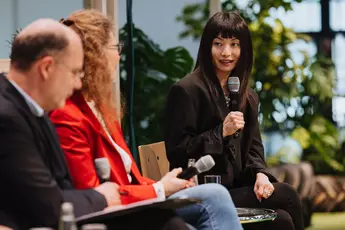 Three people holding microphones, sitting and talking in a discussion situation, plants can be seen blurred in the background.