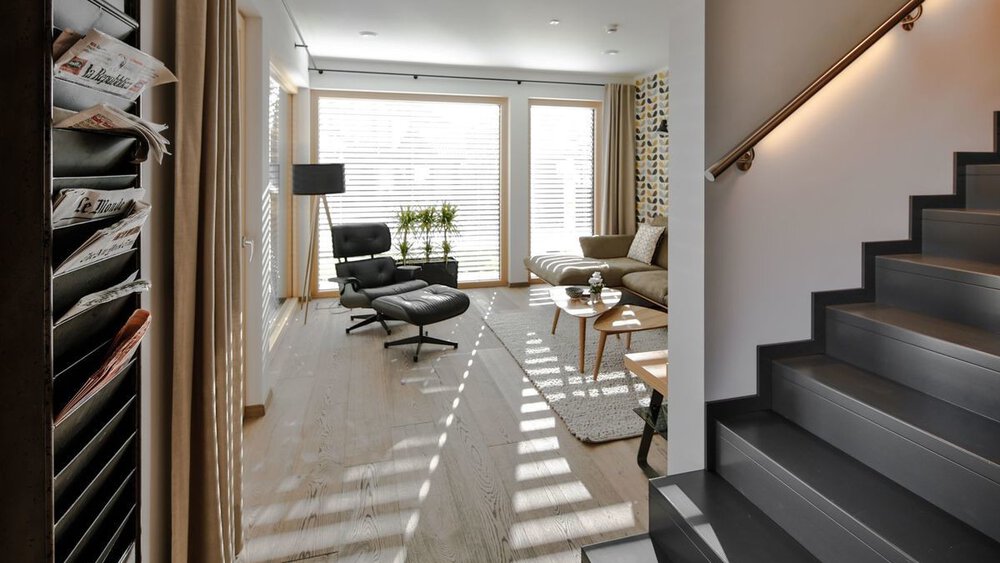 Modernes Wohnzimmer mit Loungesessel, Stehlampe und Pflanzen neben großen Fenstern und Treppe auf der rechten Seite.