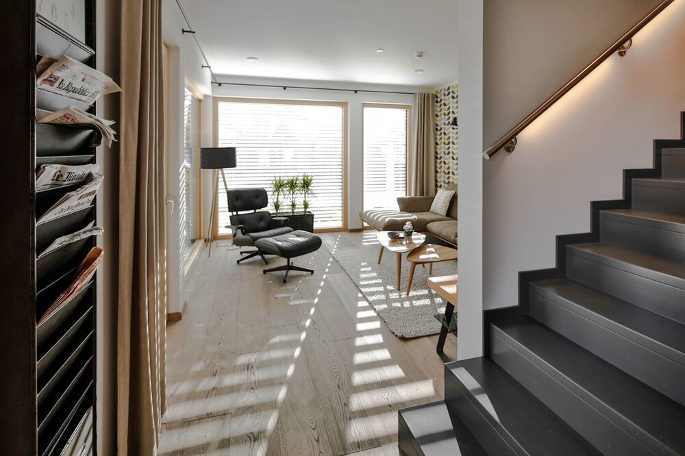 Modernes Wohnzimmer mit Loungesessel, Stehlampe und Pflanzen neben großen Fenstern und Treppe auf der rechten Seite.