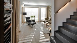 Modernes Wohnzimmer mit Loungesessel, Stehlampe und Pflanzen neben großen Fenstern und Treppe auf der rechten Seite.
