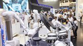 Robotic arms playing drums at a technology exhibition, people watching in the background.