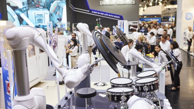 Robotic arms playing drums at a technology exhibition, people watching in the background.