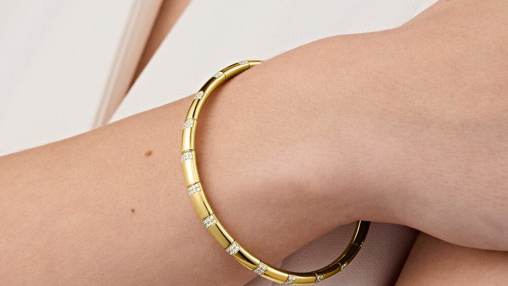 A close-up of a diamond-studded gold bracelet on the wrist of a woman wearing light-colored clothing.