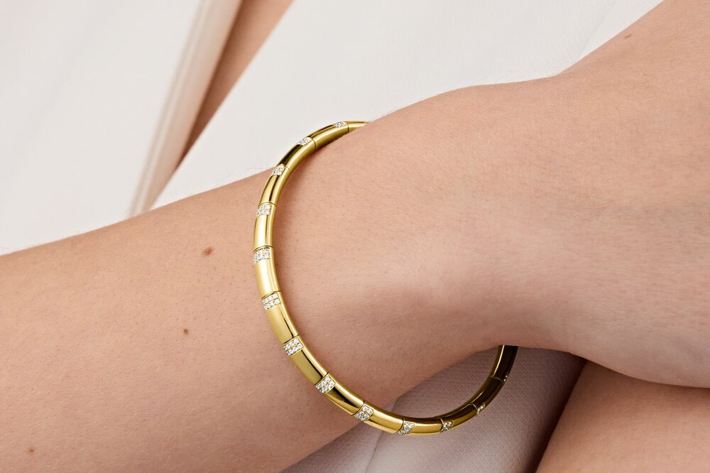 Nahaufnahme eines mit Diamanten besetzten goldenen Armreifs am Handgelenk einer Frau in heller Kleidung.