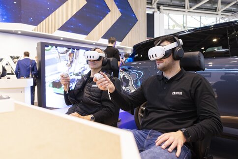 Two men sit next to each other on comfortable chairs, wearing VR headsets and holding controllers. A car is on display behind them.