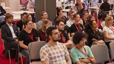 A motley group of people sit on chairs as an audience, attentively following the action on stage.