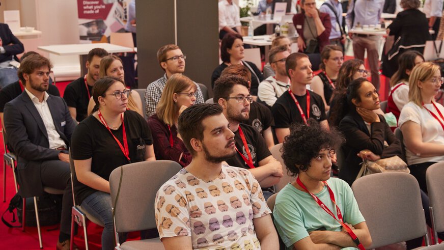A motley group of people sit on chairs as an audience, attentively following the action on stage.