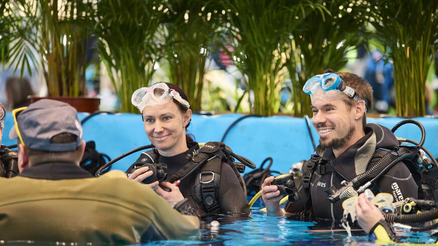 Lächelnde Taucher in Neoprenanzügen und Tauchmasken üben im Indoor-Wasserbecken der Messe f.re.e, umgeben von tropischen Pflanzen.